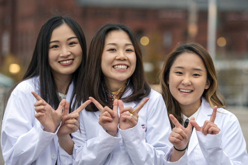 Three students wearing white coats and smiling with 'W' hands at camera after white coat ceremony