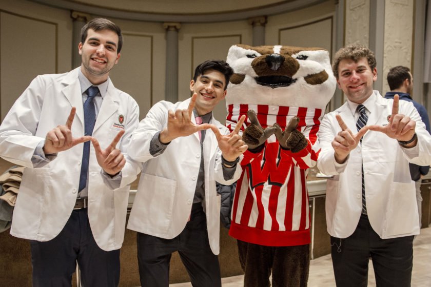 Three students wearing white coats posing with Bucky Badger and forming a 'W' with their hands, after white coat ceremony