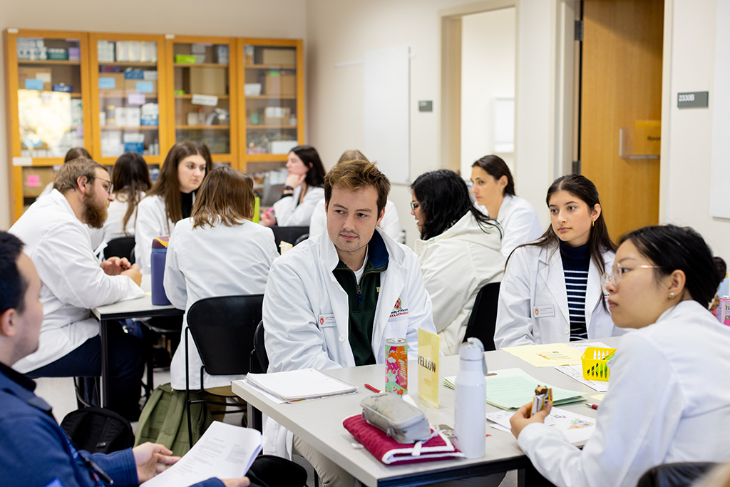 PharmD students in white coats sit at a table and listen to an instructor