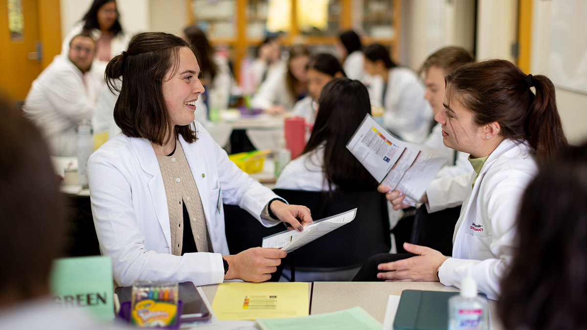Two smiling PharmD students in white coats speak facing each other while holding papers