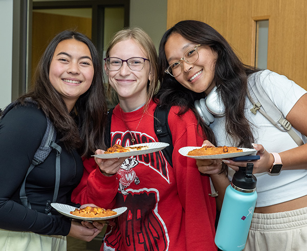 3 pharmacy students holding slices of pizza pose together with big smiles