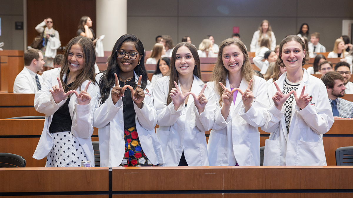 Pharmacy students in white coats holding up their hands in a W shape
