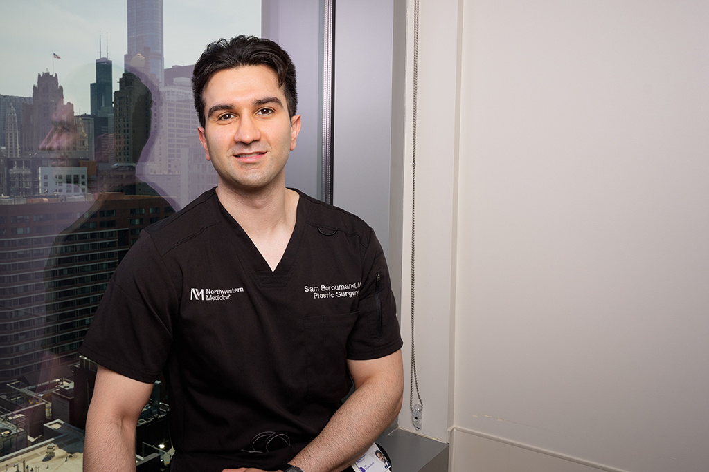 Sam Boroumand, wearing surgical scrubs, poses near a window overlooking the Chicago skyline