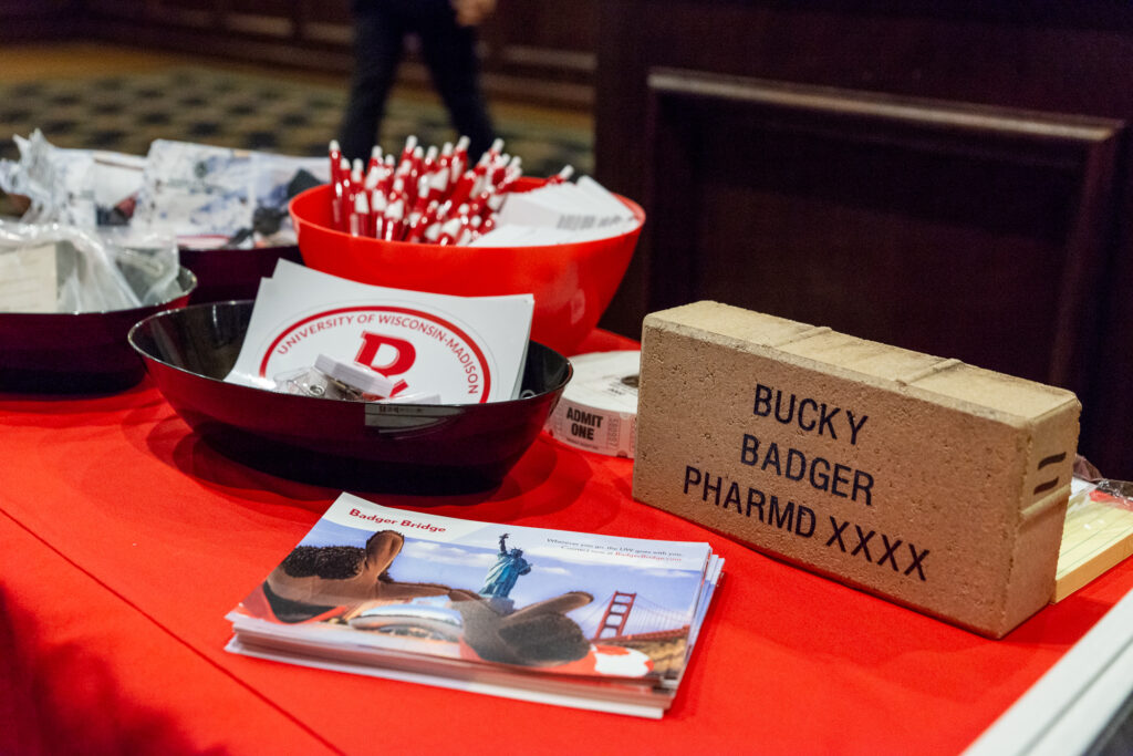 Table filled with various handouts like pens, tickets, stickers, information on Badger Bridge, and a brick labeled "Bucky Badger PharmD XXXX"