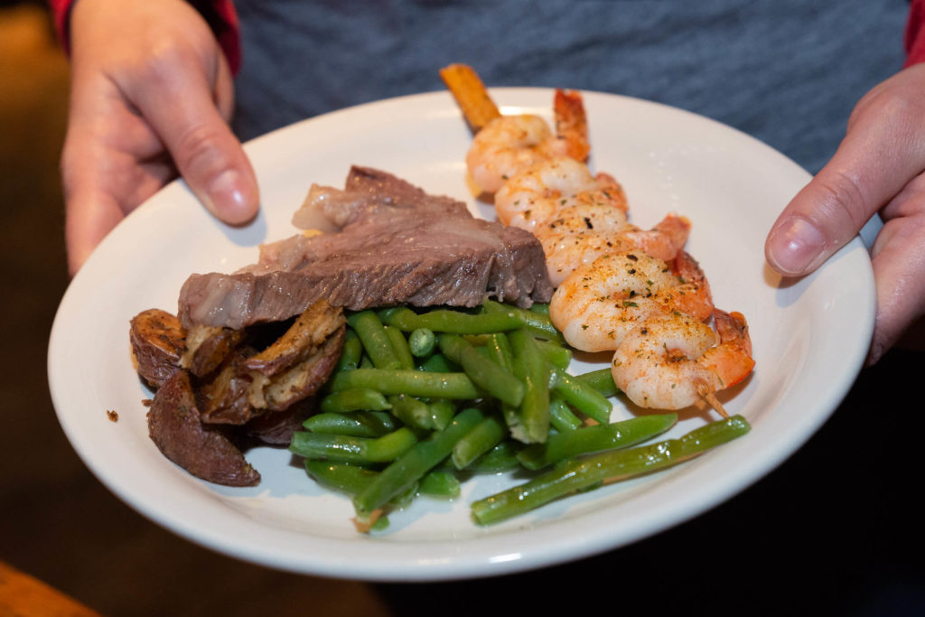 Hand holding plate of shrimp, steak, mushrooms, and green beans