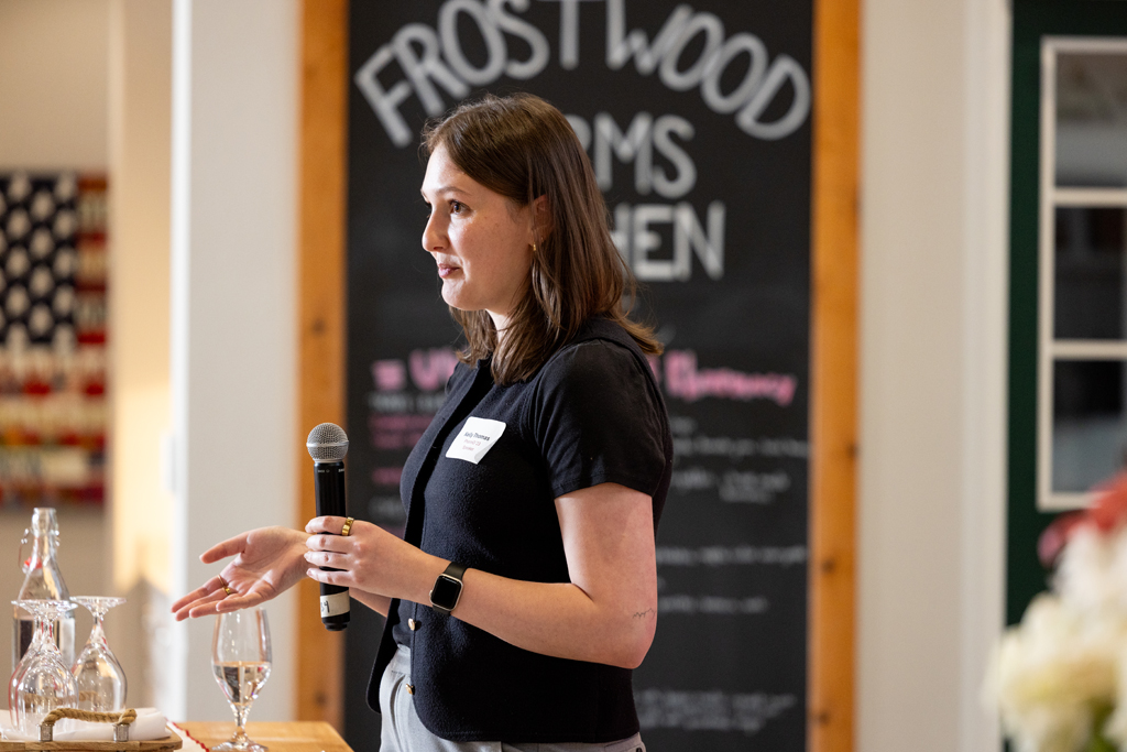 Kelly Thomas speaks into a microphone in front of a chalkboard that reads "Frostwood Farms Kitchen"