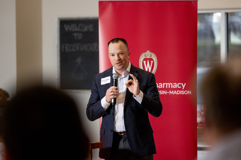 Jason Bergsbakken speaks into a microphone in front of a red School of Pharmacy banner