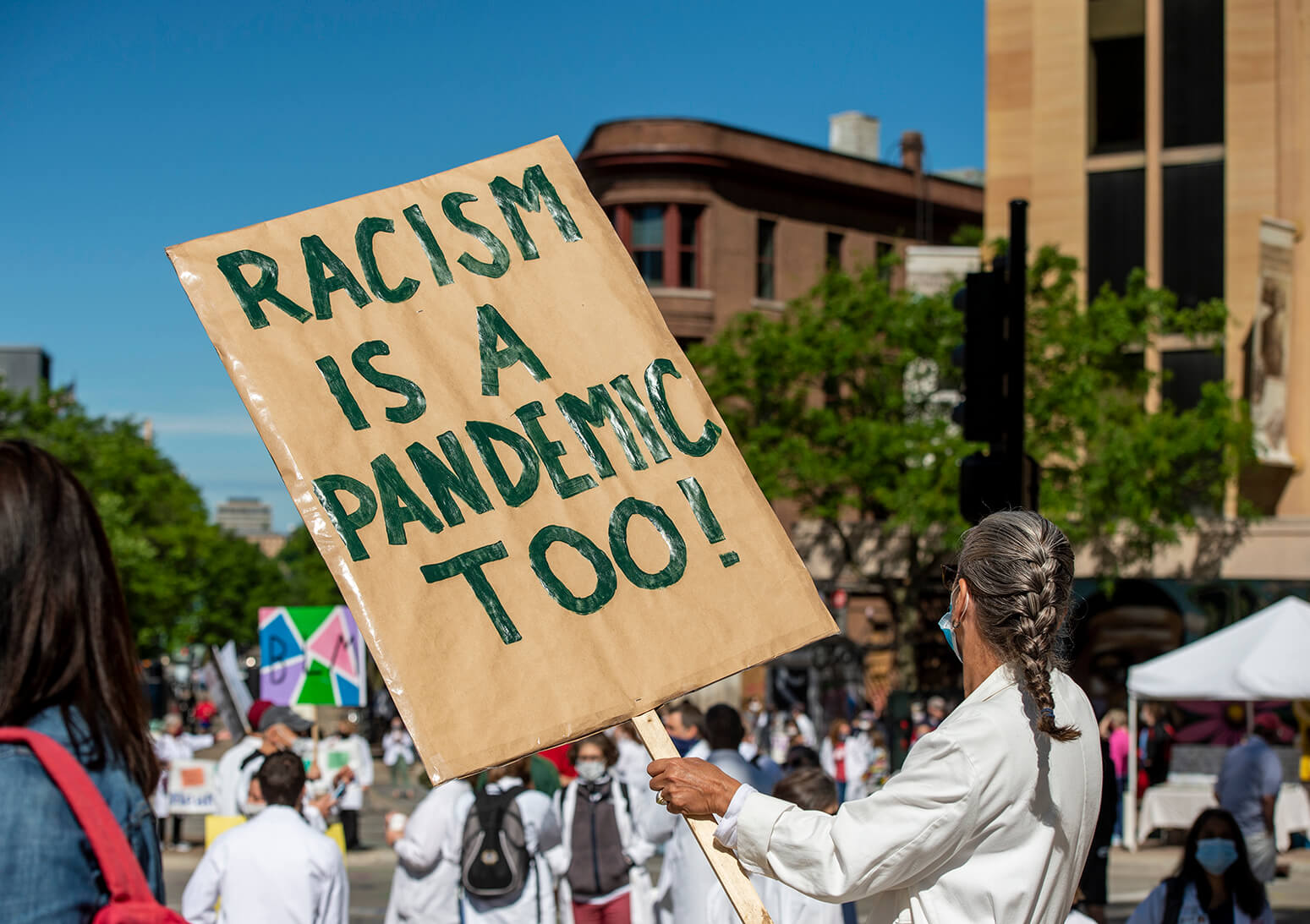 Woman in white coat and mask with sign reading "Racism is a pandemic too"