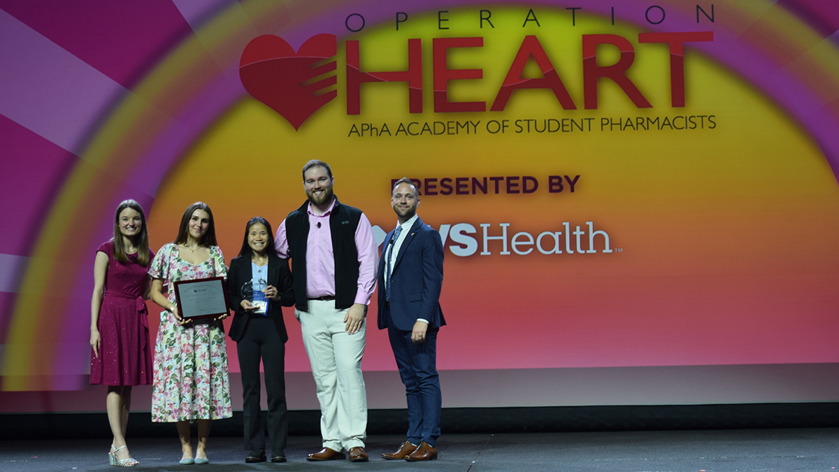 Five individuals stand onstage, with two holding awards, in front of a large screen reading Operation Heart