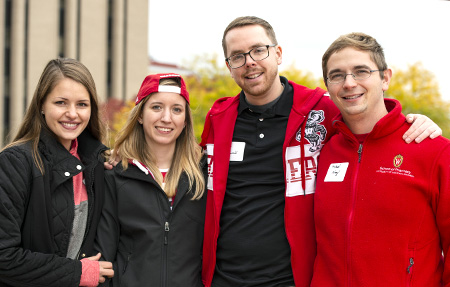 Four SoP alumni dressed in red pose together for a photo at the annual tailgate event