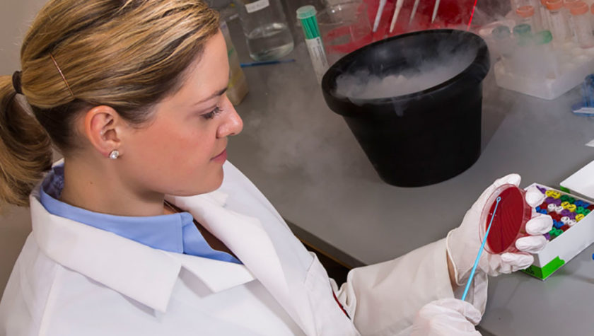 Laurel Legenza working with a substance in a petri dish and a bucket filled with smoke on the counter next to her