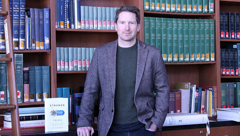 Lucas Richert next to a bookshelf with his book, Strange Trips