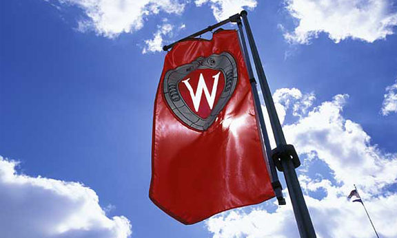 A "W" crest banner flies on Bascom Hill against blue sky and puffy clouds during spring.