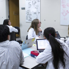 A group of PharmD students in white coats collaborating at a table in front of a white board.