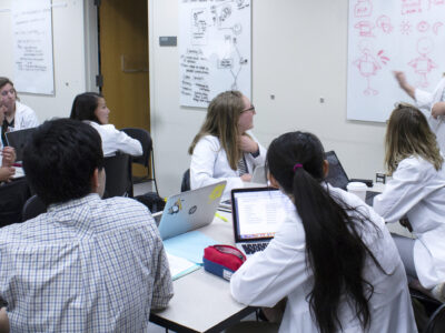 A group of PharmD students in white coats collaborating at a table in front of a white board.