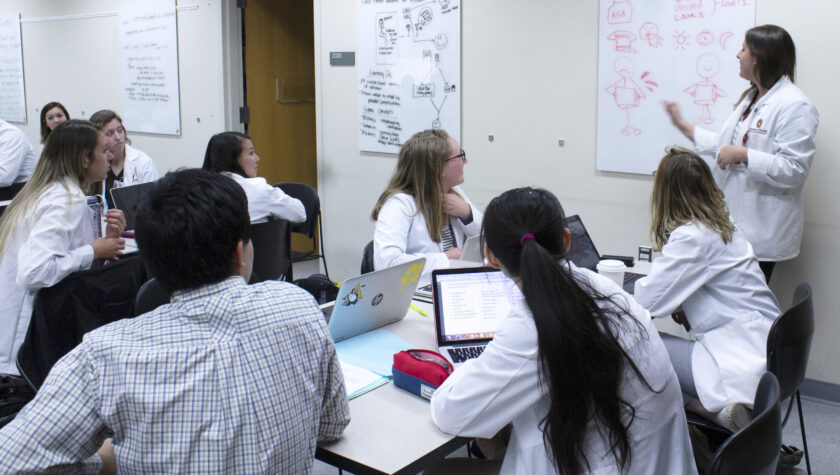 A group of PharmD students in white coats collaborating at a table in front of a white board.