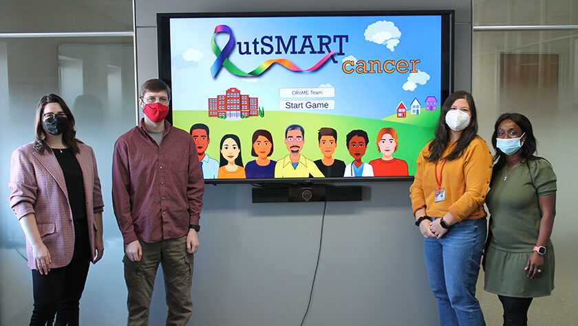 Claire Rosenberger. Eric Robinson, Lisa Szela, and Olufunmilola Abraham standing around TV displaying "outSMART cancer" game