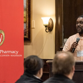 Kwadwo Owusu-Ofori speaking into a microphone in front of a UW-Madison School of Pharmacy banner