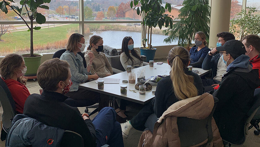 A group of students sitting around a table talking in a room full of windows.