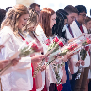 A row of PharmD students in white coats and holding red roses read from papers in their hands.