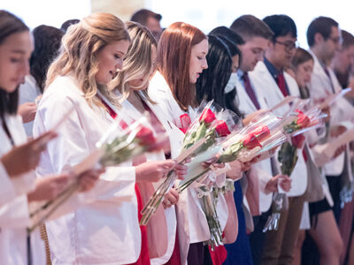 A row of PharmD students in white coats and holding red roses read from papers in their hands.