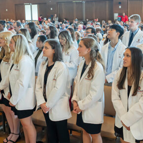 A group of students wearing white coats, looking up to read off of a screen.