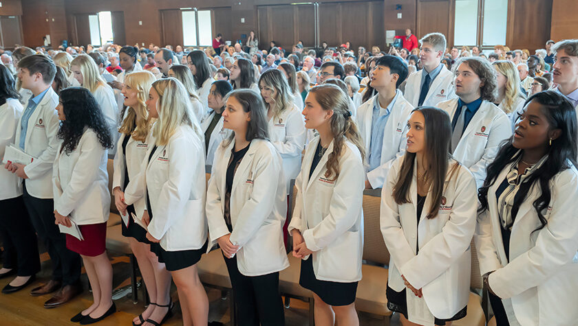 A group of students wearing white coats, looking up to read off of a screen.