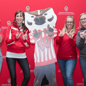 Four alumni stand with a cutout of Bucky in front of a red School of Pharmacy backdrop.