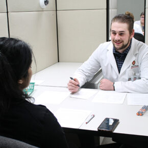 A student in a white coat speaks with a dark-haired person across a table.