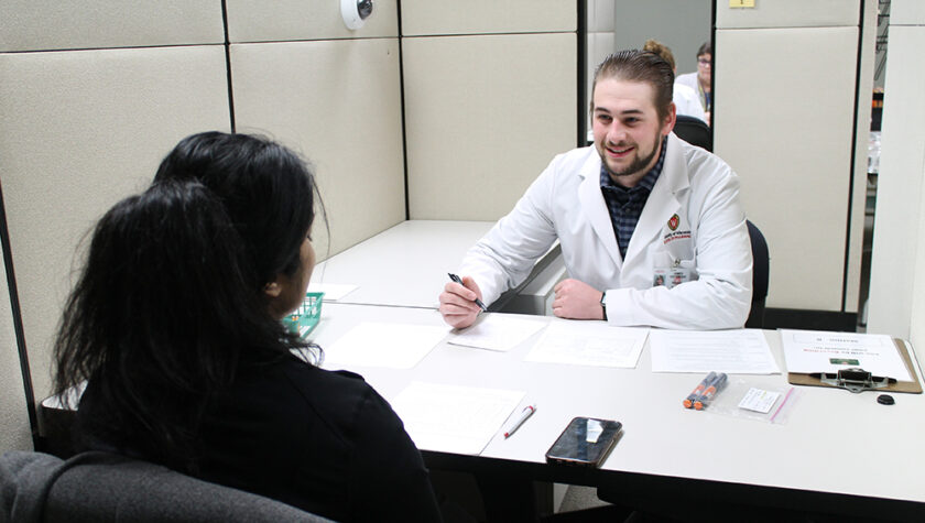 A student in a white coat speaks with a dark-haired person across a table.