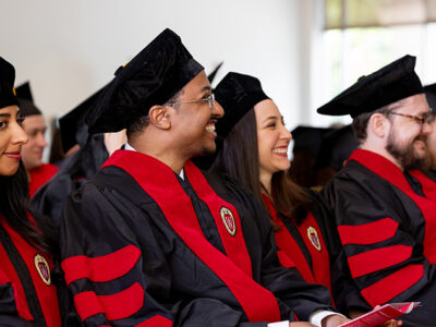 School of Pharmacy graduates in hats and robes smile while listening to a speaker.