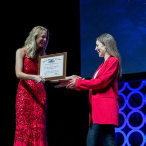 Lauren Glaza accepts an award onstage from a woman in a red dress.
