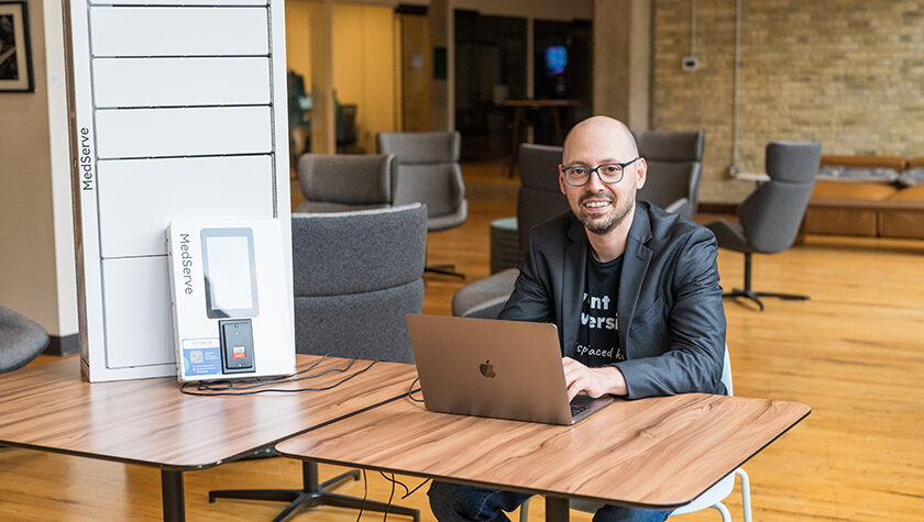 Alex Yampolsky smiles from behind a laptop, next to a white MedServe cabinet