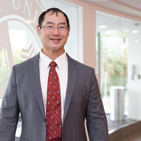 Joe Su poses in front of a window with a West Coast University logo etched into it.