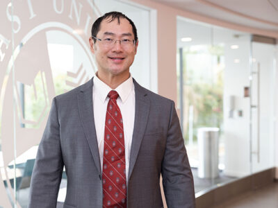 Joe Su poses in front of a window with a West Coast University logo etched into it.