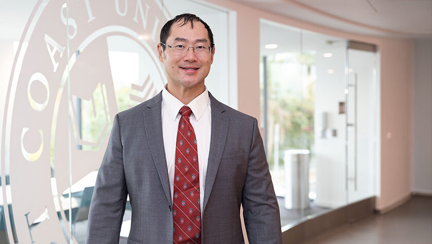 Joe Su poses in front of a window with a West Coast University logo etched into it.