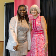 A PharmD student poses in her white coat next to Professor Mara Kieser, in a red dress.