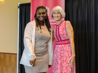 A PharmD student poses in her white coat next to Professor Mara Kieser, in a red dress.