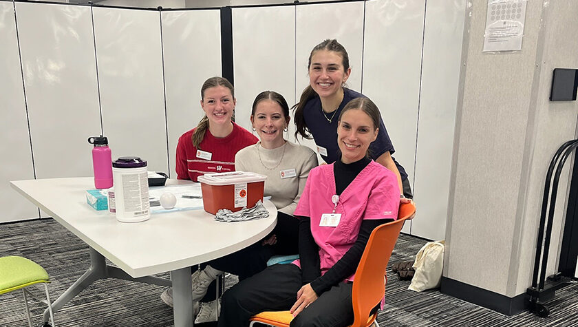 PharmD students Amalia Fox, Hannah Whitney, Chaise Pucek, and Nicole Ulbricht pose behind a white folding table with vaccination supplies on it.