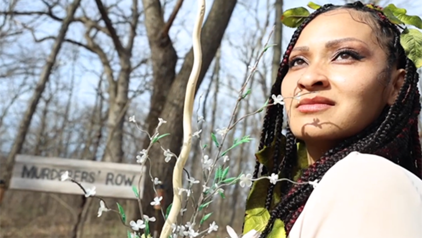 Thi Le, wearing leaves in her hair, looks back over her shoulder next to a sign reading "murderers' row."