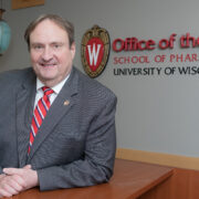 Steve Swanson poses in front of an Office of the Dean, UW–Madison School of Pharmacy sign