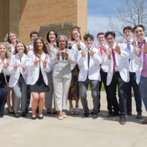 A group of PharmD students in white coats pose in front of Rennebohm Hall with W hands up