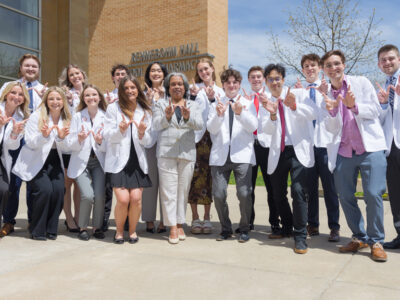 A group of PharmD students in white coats pose in front of Rennebohm Hall with W hands up
