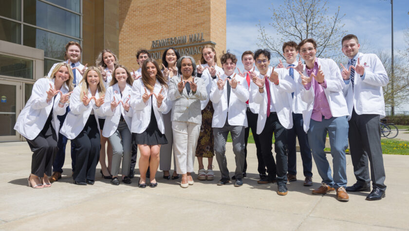 A group of PharmD students in white coats pose in front of Rennebohm Hall with W hands up