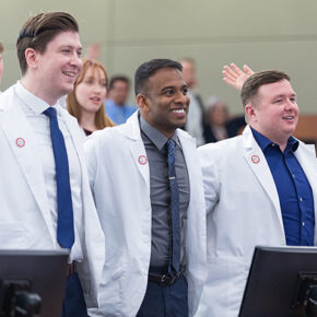 A group of PharmD students in white coats stand arm-in-arm in a lecture hall, smiling