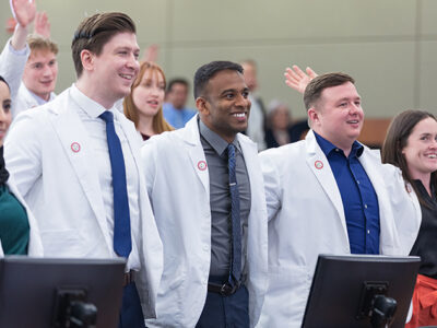 A group of PharmD students in white coats stand arm-in-arm in a lecture hall, smiling