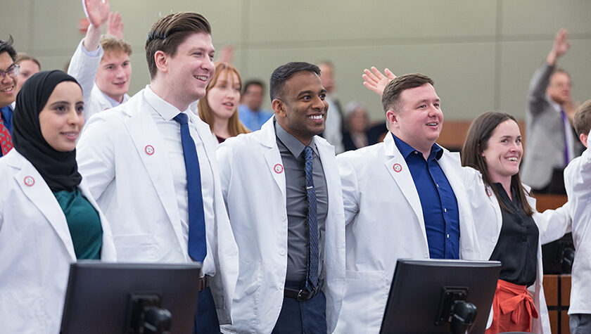 A group of PharmD students in white coats stand arm-in-arm in a lecture hall, smiling