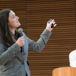 Jenna Vandehey holds a microphone and a small black cylinder while gesturing next to her