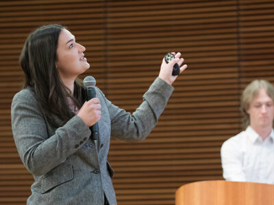 Jenna Vandehey holds a microphone and a small black cylinder while gesturing next to her