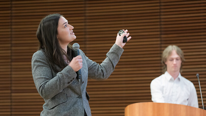 Jenna Vandehey holds a microphone and a small black cylinder while gesturing next to her